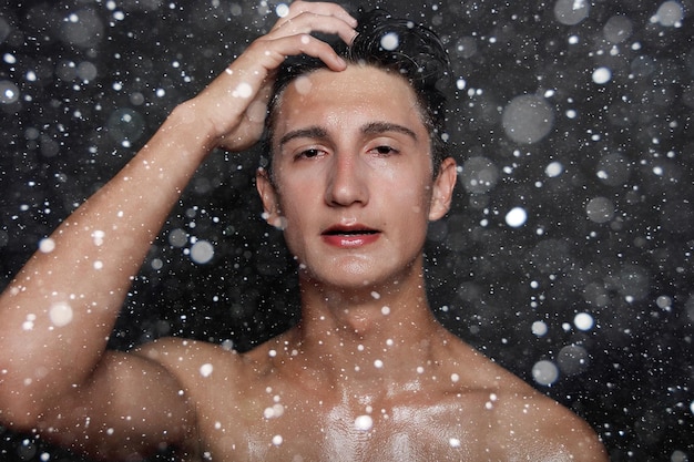 Conceito de neve, inverno, natal, pessoas, skincare e beleza - jovem molhado com cabelo preto em um fundo preto de neve. retrato masculino com peito raspado. cuidados com a pele dos homens.