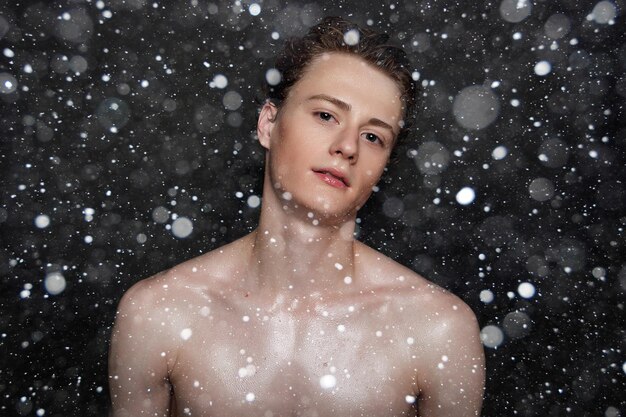 Conceito de neve, inverno, Natal, pessoas, skincare e beleza - jovem molhado com cabelo preto em um fundo preto de neve. Retrato masculino com peito raspado. Cuidados com a pele dos homens.