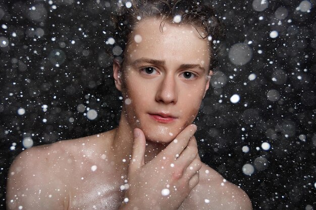 Conceito de neve, inverno, Natal, pessoas, skincare e beleza - jovem molhado com cabelo preto em um fundo preto de neve. Retrato masculino com peito raspado. Cuidados com a pele dos homens.