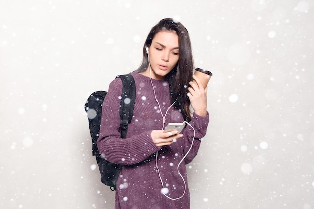 Conceito de neve, inverno, Natal, tecnologia, estilo de vida, vício em internet e pessoas - jovem mulher bonita com telefone inteligente. mulher sorridente e atraente olhando para o fundo de neve do celular