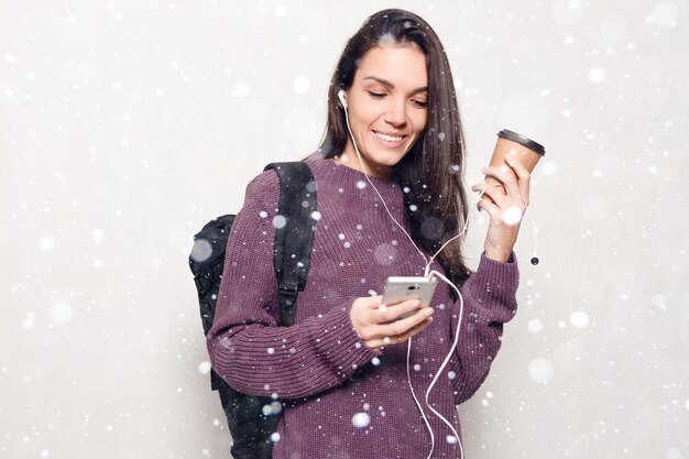 Conceito de neve, inverno, Natal, tecnologia, estilo de vida, vício em internet e pessoas - jovem mulher bonita com telefone inteligente. mulher sorridente e atraente olhando para o fundo de neve do celular