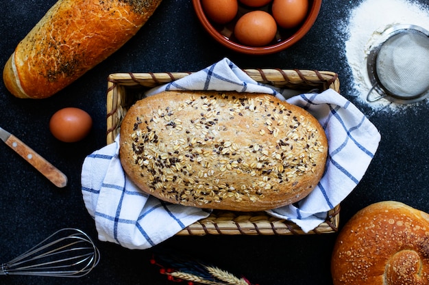 Conceito de padaria. Uma variedade de tipos diferentes de pão em um fundo escuro. Pão em uma cesta, ovos, farinha, espigas de trigo. Vista superior (postura plana). Espaço para texto