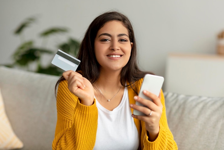 Foto conceito de pagamento online senhora muçulmana sorridente segurando cartão de crédito e smartphone sentado no sofá e olhando para a câmera