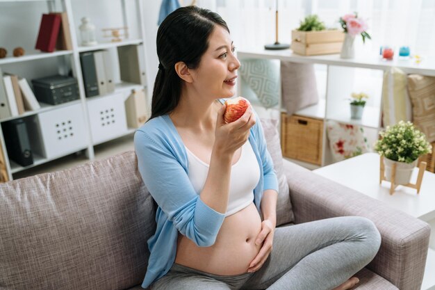conceito de paternidade de maternidade de beleza de cuidados de saúde. bela jovem chinesa grávida segurando uma maçã vermelha comer sorrindo alegria olhar janela durante o dia na sala de estar no sofá. mão feminina toque na barriga