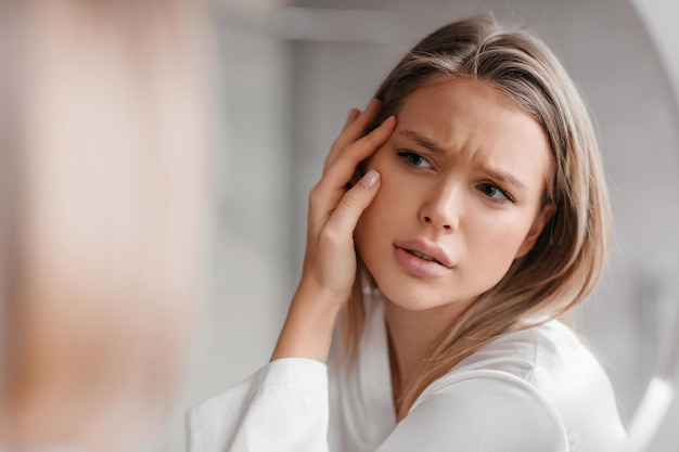 Conceito de pele propensa à acne senhora triste olhando para o espelho e verificando a pele do rosto em pé no banheiro