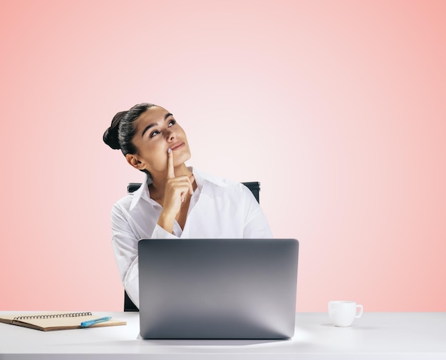 Conceito de pensamento com jovem pensativa olhando para cima e tentando encontrar a melhor solução sentado na frente do laptop moderno isolado no fundo da parede rosa claro closeup