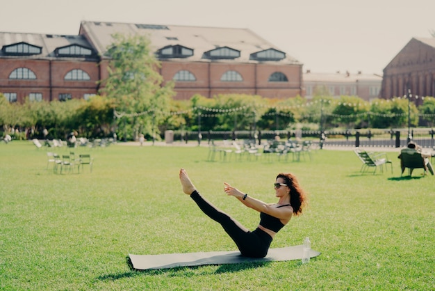 Foto conceito de perda de peso e fitness de pilates. mulher jovem morena positiva tem treinamento ao ar livre levanta poses de pernas no treino regular de karemathas usa roupas esportivas, óculos de sol, alonga os músculos.