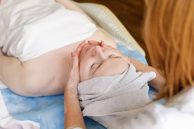 Conceito de pessoas, beleza, spa, cosmetologia e tecnologia - Mulher jovem e bonita recebendo massagem facial com os olhos fechados em um salão de spa