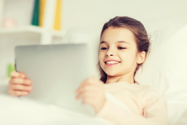 conceito de pessoas, crianças, descanso e tecnologia - garota sorridente feliz deitada acordada com computador tablet pc na cama em casa