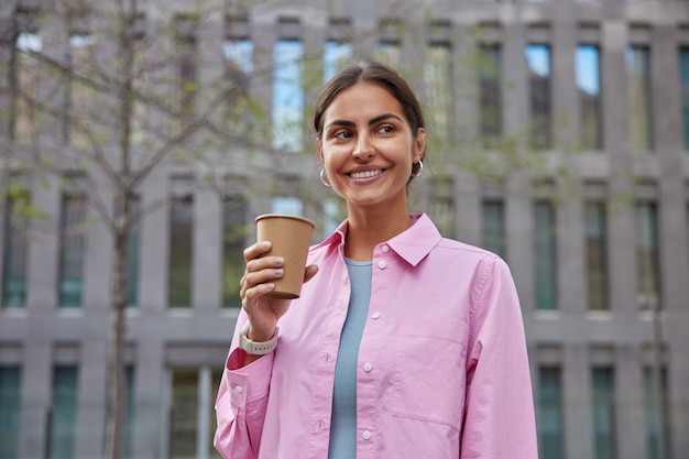 Conceito de pessoas e lazer. jovem positiva passeia pela cidade faz excursão a pé, pausa para o trabalho, aproveita o fim de semana e clima de primavera bebe café para viagem usa camiseta rosa posa perto do prédio