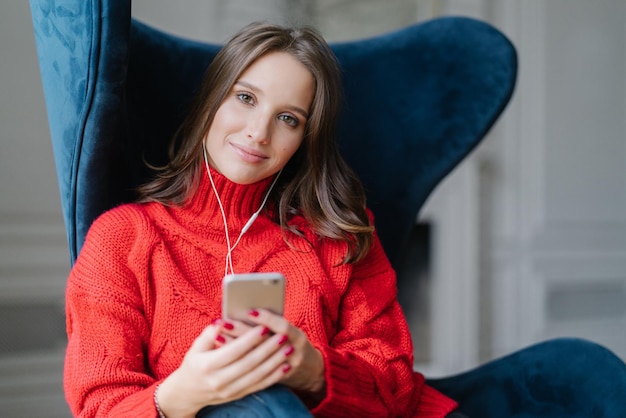 Conceito de pessoas e tecnologia meloman feminino satisfeito gosta de música favorita ouve registro de áudio vestido com suéter vermelho surfa na lista de reprodução senta-se na poltrona na sala tem tempo livre