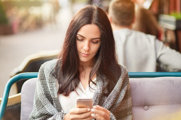 Conceito de pessoas, estilo de vida e tempo livre. Jovem mulher atraente possui telefone celular moderno, assiste o vídeo através do aplicativo de mídia, embrulhado em xadrez