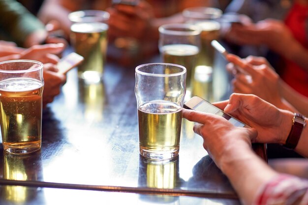 conceito de pessoas, lazer, tecnologia e bebidas - amigos bebendo cerveja e mensagens de texto em smartphones no bar ou pub