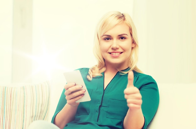 conceito de pessoas, tecnologia, comunicação e lazer - jovem feliz sentada no sofá com smartphone em casa e mostrando polegares para cima