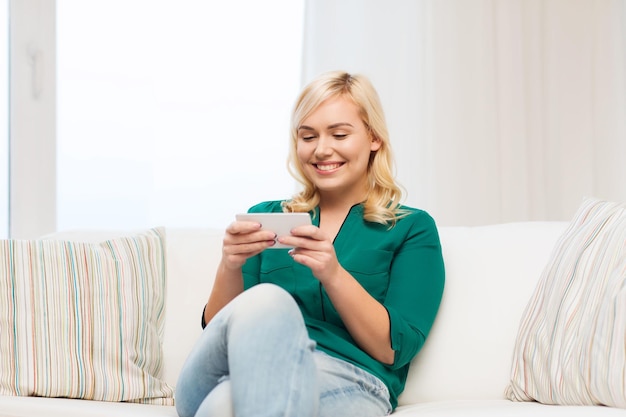 conceito de pessoas, tecnologia, comunicação e lazer - jovem feliz sentada no sofá com smartphone em casa