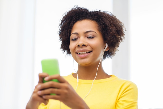 Conceito de pessoas, tecnologia e lazer - feliz jovem afro-americana sentada no sofá com smartphone e fones de ouvido ouvindo música em casa