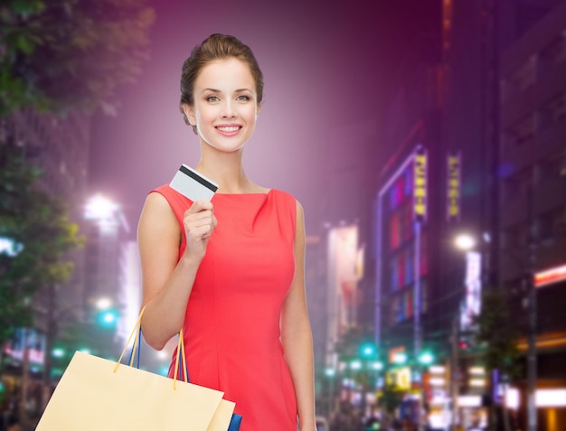 conceito de pessoas, venda, natal e feriados - sorridente mulher elegante de vestido vermelho com sacolas de compras e cartão de crédito sobre o fundo da cidade à noite