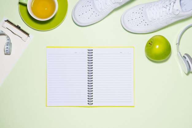 Conceito de plano de fitness. Tênis, chá, maçã e fone de ouvido no fundo da cor pastel com o caderno aberto.
