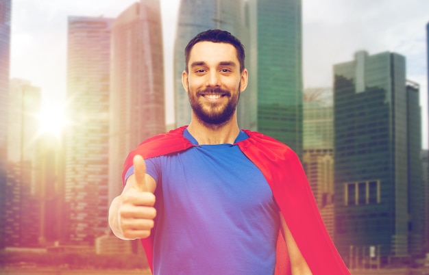 Foto conceito de poder e pessoas - homem feliz em capa vermelha de super-herói mostrando polegares para cima sobre fundo de arranha-céus da cidade de singapura