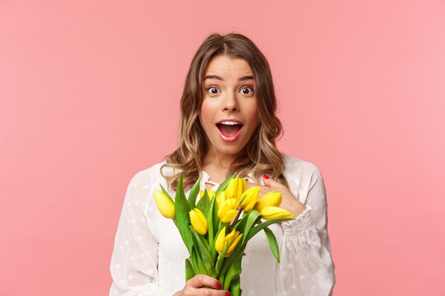 Conceito de primavera, felicidade e celebração. O close-up da menina loura nova surpreendida e maravilhada no vestido branco, recebe tulipas amarelas e apontando-se com descrença e espanto, não exatos