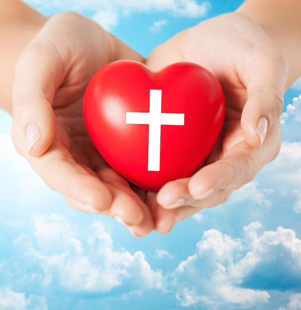 conceito de religião, cristianismo e caridade - close-up de mãos femininas segurando um coração vermelho com símbolo de cruz cristã sobre fundo de céu azul e nuvens