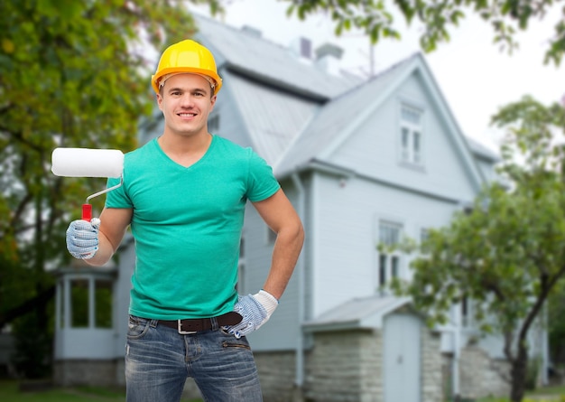 conceito de reparação, construção, pessoas, construção e manutenção - trabalhador manual masculino sorridente no capacete protetor segurando o rolo de pintura sobre o fundo da casa