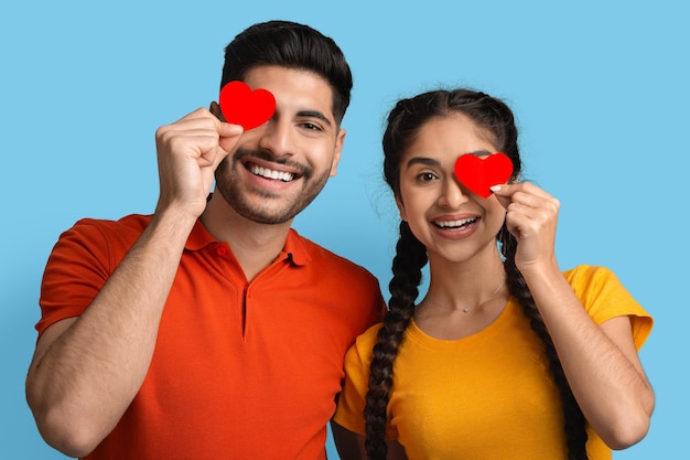 Conceito de romance amoroso casal árabe segurando corações vermelhos perto dos olhos e sorrindo