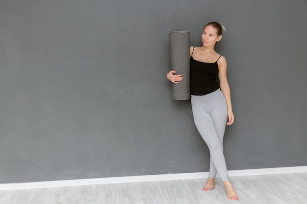 Conceito de rotina de treino Mulher magra feliz segurando o tapete de ioga na mão em pé contra a parede