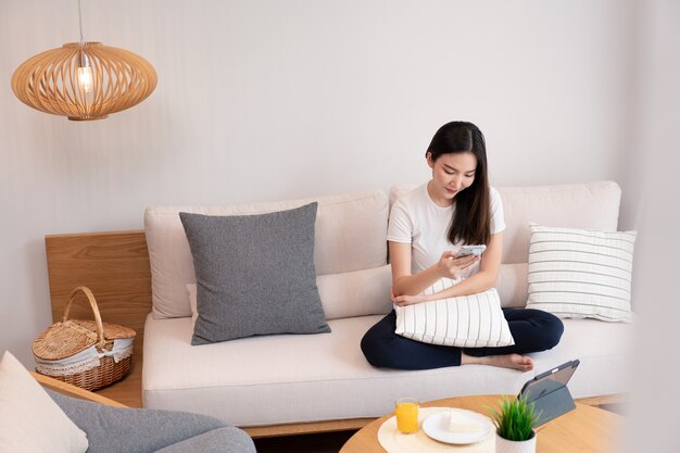 Conceito de sala de estar uma menina bonita sentada no sofá confortável, gastando seu lazer on-line com suco e lanches na sala de estar.