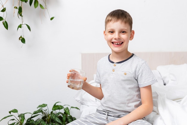 Conceito de saúde Criança ativa feliz sorrindo quando segura água pura e fresca em um copo na cama pela manhã Cuidando do seu corpo Ritual de Despertar