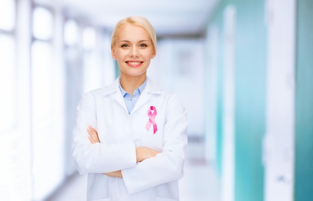 conceito de saúde e medicina - médica sorridente com fita rosa de conscientização do câncer sobre fundo hospitalar