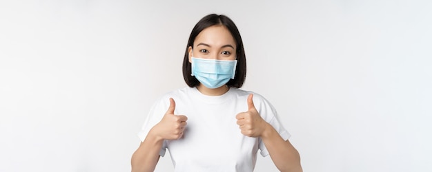 Conceito de saúde e pessoas Covid19 Jovem coreana feliz em máscara médica mostrando os polegares para cima em camiseta sobre fundo branco