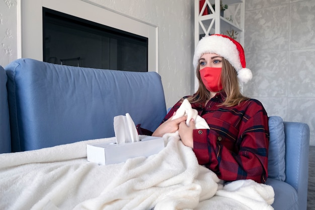 Foto conceito de saúde, frio, férias e pessoas. mulher jovem doente e triste com máscara médica em casa. jovem loira de camisa vermelha e chapéu de natal deitada no sofá com guardanapos