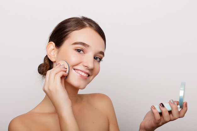 Conceito de saúde, pessoas e beleza - jovem aplicando blush no rosto com esponja de pó, conceito de cuidados com a pele, composição da foto de menina morena, isolado no fundo branco