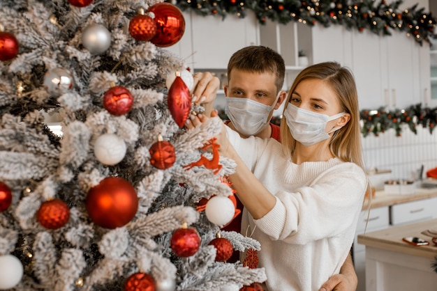 Conceito de saúde, pessoas e férias de inverno - casal usando máscara médica protetora para se proteger do coronavírus está decorando a árvore de Natal em casa