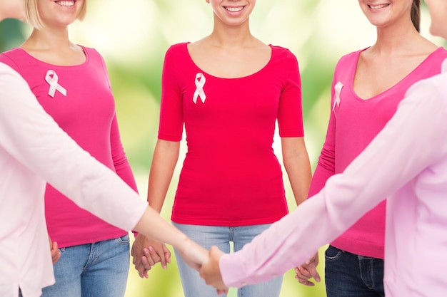 conceito de saúde, pessoas e medicina - close-up de mulheres sorridentes em camisas em branco com fitas de conscientização de câncer de mama rosa segurando as mãos sobre fundo verde