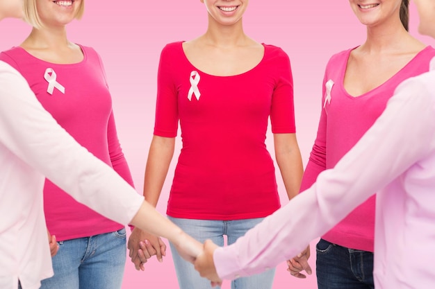 Conceito de saúde, pessoas e medicina - close-up de mulheres sorridentes em camisas em branco com fitas de conscientização de câncer de mama segurando as mãos sobre fundo rosa