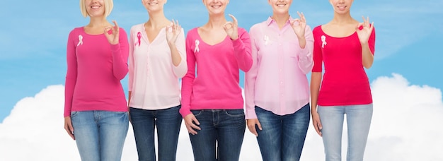 conceito de saúde, pessoas, gesto e medicina - close-up de mulheres sorridentes em camisas em branco com fitas de conscientização de câncer de mama rosa mostrando sinal de ok sobre céu azul e fundo de nuvem branca