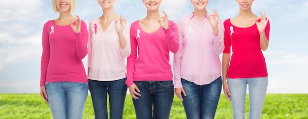 conceito de saúde, pessoas, gesto e medicina - close-up de mulheres sorridentes em camisas em branco com fitas de conscientização de câncer de mama rosa mostrando sinal de ok sobre fundo de céu azul e grama