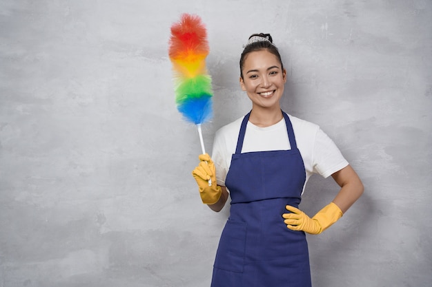 Conceito de serviço de arrumação e limpeza. Mulher jovem feliz, faxineira de uniforme segurando um espanador de microfibra colorido e sorrindo para a câmera em pé contra a parede cinza