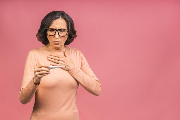 Conceito de sintoma do coronavírus. Mulher idosa madura com gripe, segurando um termômetro eletrônico na mão, isolada em um fundo rosa com espaço de cópia