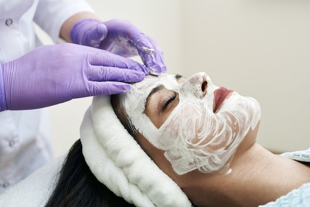 Conceito de spa. Jovem mulher com máscara facial de nutrientes em salão de beleza, close-up. A esteticista aplica uma máscara hidratante no rosto da cliente.
