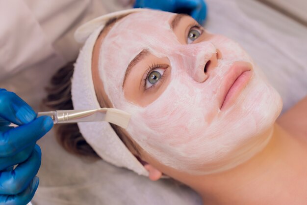 Foto conceito de spa. jovem mulher com máscara facial de nutrientes em salão de beleza, close-up. a esteticista aplica uma máscara hidratante no rosto da cliente.