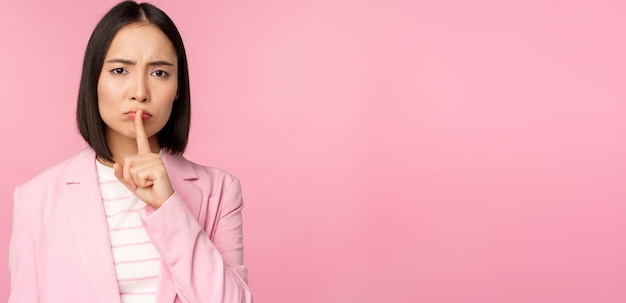 Conceito de tabu de silêncio Retrato de empresária asiática mostrando gesto de silêncio shhh sinal pressione o dedo nos lábios em pé sobre fundo rosa de terno