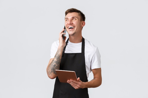 Conceito de take-away de lojas de varejo, pequenas empresas, cafés e restaurantes. Lindo vendedor sorridente, barista falando ao telefone, rindo feliz, anotando pedido para entrega, segurando tablet digital