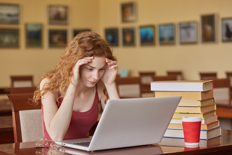 Conceito de tarefa, teste, aprendizagem e educação. Inteligente, garota leitor vestindo casualmente sentado na mesa na sala de leitura na frente do laptop, mantém as mãos na testa, preparando-se para um exame importante.