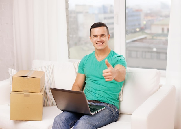 Foto conceito de tecnologia, casa e estilo de vida - homem sorridente com laptop e caixas de papelão em casa mostrando os polegares