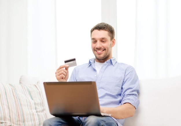 conceito de tecnologia, casa e estilo de vida - homem sorridente trabalhando com laptop e cartão de crédito em casa