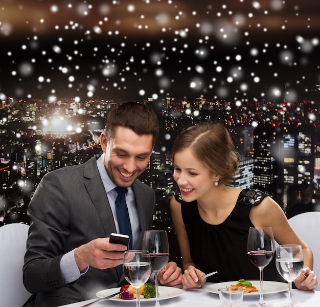 conceito de tecnologia, comida, natal, feriados e pessoas - casal sorridente com smartphone comendo no restaurante sobre o fundo da cidade de noite de neve