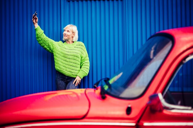 Conceito de tecnologia de pessoas felizes do telefone móvel selfie com linda loira com roupas verdes e carro vintage vermelho e parede azul. pessoa milenar moderna, estilo de vida moderno e independência